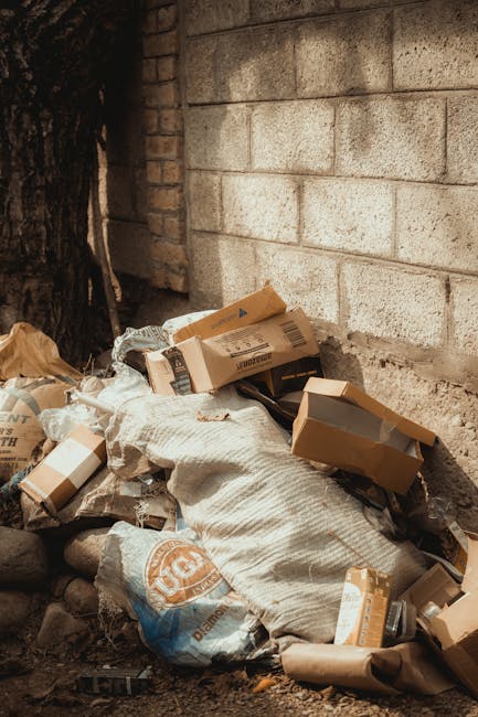 A pile of mixed waste materials situated outdoors against a weathered brick and concrete wall. The refuse includes multiple flattened cardboard boxes of various sizes, some with visible printed labels, and crumpled brown paper, all scattered haphazardly on the ground. A large, textured, off-white sack made of woven fabric partially covers the debris, with parts of the cardboard boxes resting on and around it. The surrounding environment appears to be an alleyway or side of a building, with a dark tree trunk partly visible on the left side. The ground is uneven, comprised of dirt and small stones, indicating an informal rubbish collection area. The scene depicts typical private waste disposal activity, similar to those handled by independent rubbish removal services such as Rubbish Removal Knightsbridge, emphasizing proper compacting and clearing of waste materials from urban or residential settings. The lighting is natural but subdued, fitting an outdoor setting during the daytime, with no visible equipment or vehicles in the scene.