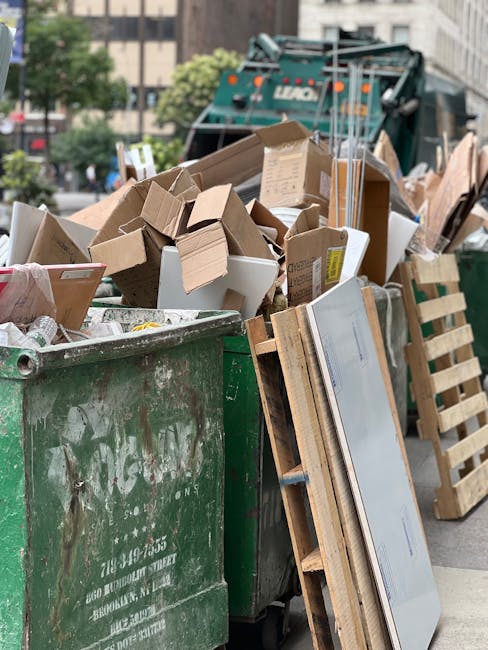 A large green waste skip filled with various types of rubbish, including numerous broken cardboard boxes, some flattened and others partially assembled, with visible creases and corrugated textures. The skip is situated outdoors on a paved urban street, showing signs of weathering with rust and dirt marks on its surface. Leaning against the side of the skip is a wooden pallet with rough, unfinished texture and light brown color, partially obscured by the rubbish. In the background, a compact garbage collection truck is visible, predominantly dark green with orange safety lights on top, and is parked near multi-storey buildings with windows and balconies, indicating a residential or commercial area. The scene is illuminated by natural daylight, giving a clear view of the clutter and suggesting an ongoing rubbish removal operation. This image exemplifies private waste management or on-site clearance, typical of services like those offered by Rubbish Removal Knightsbridge, providing an alternative to council-led rubbish collection in busy urban environments.