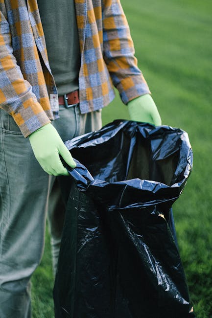 A person standing outdoors on a grassy area, holding open a large black rubbish bag with their hands gloved in green. The individual is dressed in casual clothing, including a blue shirt, a yellow and blue plaid jacket, and light-colored trousers. The black rubbish bag is made of thick plastic, lined with shiny material, and appears to be empty or ready for waste collection. The background features a well-maintained lawn, suggesting the scene is set in a garden or park environment. This image relates to private waste handling or rubbish removal services, such as those offered by Rubbish Removal Knightsbridge, highlighting actions involved in on-site clearance or rubbish collection outside of traditional municipal disposal. The natural lighting emphasizes the textures of the gloves, clothing, and the glossy surface of the rubbish bag, reflecting a professional yet neutral scene focused on waste collection preparation.
