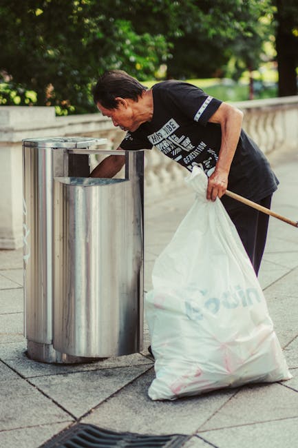 A middle-aged man with dark hair, wearing a black T-shirt with white text and dark trousers, is seen disposing of rubbish into a large, cylindrical stainless steel public trash bin situated on a paved sidewalk. His right hand is reaching into the open top of the bin, while his left hand holds a white plastic bag filled with waste, which rests against his leg. The trash bin is set against a low stone railing, with a background of lush green foliage and trees, suggesting a park or urban green space. The scene is illuminated by natural daylight, casting soft shadows, and the environment appears clean and well-maintained. This setting illustrates an example of private or independent waste disposal, similar to rubbish removal services, as the individual manages waste outside of local authority collection systems, aligning with the context of rubbish clearance options in a professional waste management scenario.