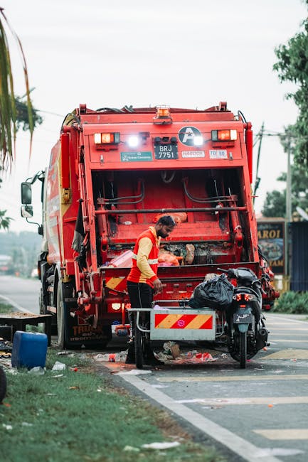 In a roadside setting, a worker wearing a high-visibility yellow and red vest is operating a red rubbish collection truck, which is positioned on the pavement. The back of the truck is open, revealing mechanical arms and storage compartments used for waste collection. The worker is mid-action, possibly sorting or managing rubbish, with a small black waste bag placed on a nearby platform attached to a motorbike parked alongside the truck. Surrounding the scene, there are patches of grass and a sidewalk, with a few other vehicles and trees in the background, indicating an urban or suburban environment. Overhead, the sky appears overcast, providing diffuse natural lighting that highlights the textures of the truck’s metal surfaces, the worker’s attire, and the scattered rubbish on the ground. This scene exemplifies independent waste collection or on-site rubbish disposal, aligning with private rubbish removal services rather than municipal collection, and emphasizes the practical aspects of waste management in a community context.