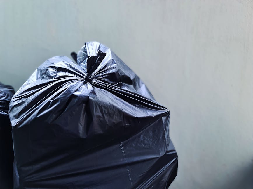 A pile of discarded rubbish situated on a gravel surface next to a stone wall and a metal pole. The waste includes black plastic rubbish bags, some partially torn open revealing miscellaneous waste inside. There is a large, beige, worn-out car tire leaning against the pile, revealing visible tread and sidewall details. A bright yellow plastic container lies partially beneath the debris, contrasting with the dark bags. In the background, overgrown bushes extend along the top of the stone wall, with a metal fence and a large, curved, semi-permanent shelter structure visible behind. Overhead, electrical wires extend across a partly cloudy sky, suggesting an outdoor setting possibly near a waste collection or disposal site. The scene captures a typical scenario of unmanaged waste accumulation that private rubbish removal services, such as those offered by Rubbish Removal Knightsbridge, might address through on-site clearance or alternative waste handling solutions.