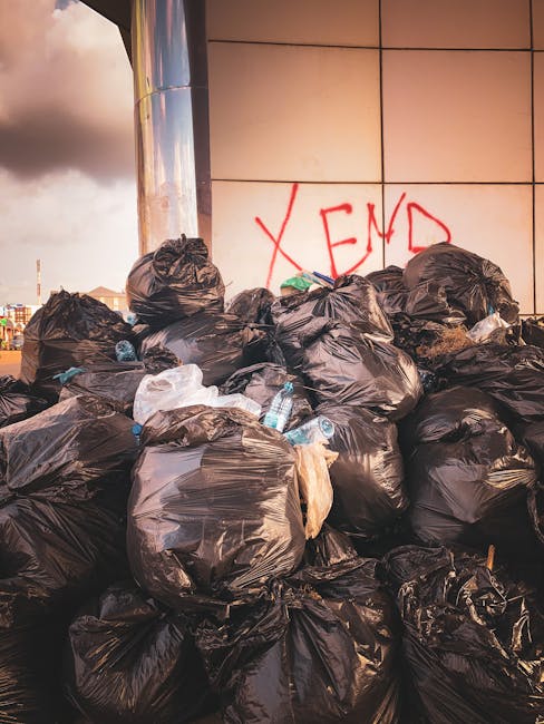 A large pile of black garbage bags is stacked against a tiled wall of modern building exterior, some bags appearing full and tightly sealed while others are slightly torn or crumpled. Several clear plastic bottles are visibly protruding from the bags, indicating mixed waste. Behind the bags, on the wall, there is red graffiti spelling 'XEND'. The setting suggests an urban environment, possibly an alley or service area, with the piles of waste indicating an instance of unmanaged or independent rubbish disposal. This scene aligns with scenarios where private waste collection services may be required, reflecting the context of alternative rubbish removal options that Rubbish Removal Knightsbridge offers for non-council supported waste clearance.