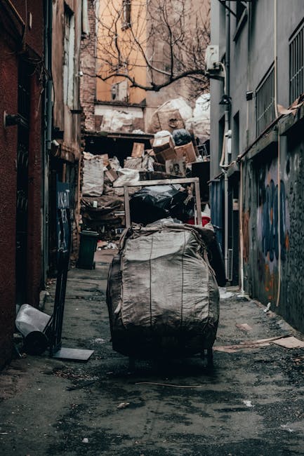 A pile of mixed rubbish, including cardboard boxes, paper bags, and plastic packaging, is positioned on the ground against a rough, weathered brick wall and a concrete block walltle. The cardboard boxes vary in size, some with visible flaps, and are made of plain brown material with printed labels and barcodes. There are also plastic and paper bags, some with printed logos, and a large white and orange sack partially visible underneath the rubbish. The debris is loosely arranged, with some boxes leaning or partially opened, revealing shadows within. The surrounding environment appears to be an outdoor area, such as a back alley or side yard, with gravel and small stones scattered at the base of the wall. A vertical section of tree trunk or thick branch is visible in the background to the left. The scene is illuminated with natural light, creating soft shadows and highlighting the textures and materials of the waste, which is indicative of an area designated for uncollected or privately disposed rubbish, relevant to independent clearance services. Rubbish Removal Knightsbridge may handle such waste removal tasks, including on-site clearance of domestic or commercial debris.