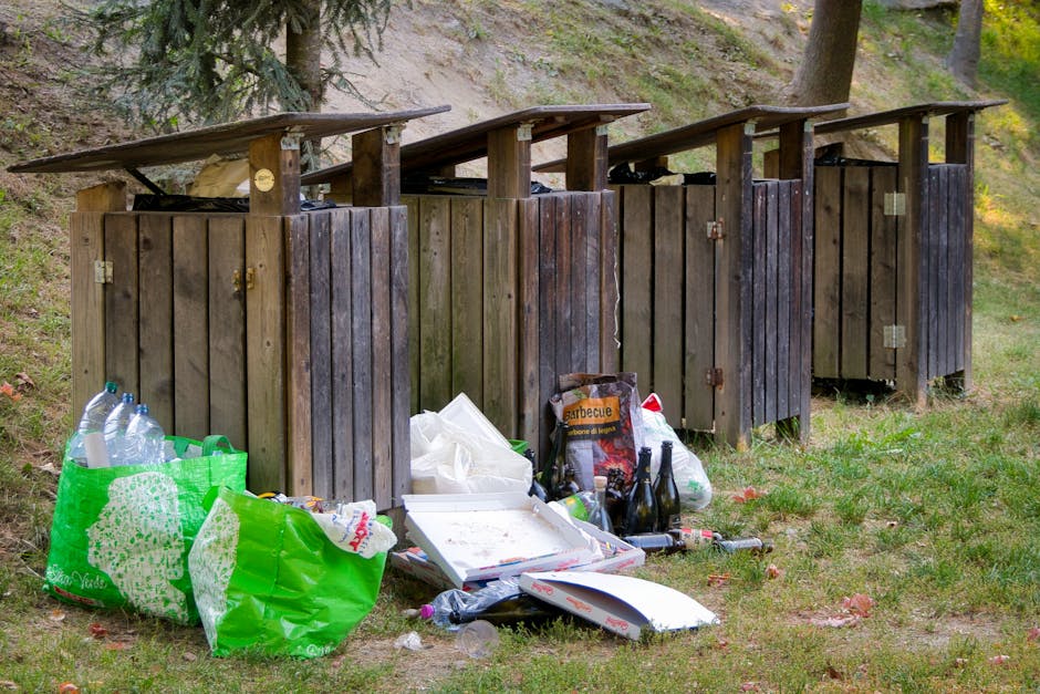 A row of four dark brown wooden outdoor rubbish bins with sloped roofs arranged on a grassy area next to a slope with trees in the background. The bins are weathered, with some visible hinges and locks. In front of the bins, there are discarded items including a green plastic bag filled with empty bottles and waste, a white pizza box with food remnants, and several empty glass bottles. The scene appears to be an outdoor waste disposal area, potentially related to private rubbish collection services, as it contrasts with official council rubbish disposal methods. The environment is natural with grass and trees, and the overall lighting suggests daylight with some shadows cast by the trees.