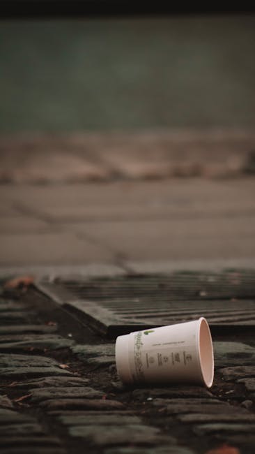 A white paper cup with printed text lies on its side on a weathered brick pavement, near a metal drainage grate. The scene appears to be outdoors, with a blurred background of a concrete or stone surface and possibly a grassy or mossy area beyond. The lighting suggests natural daylight, casting soft shadows. The cup's surface is smooth with writing that is partially visible but not legible, indicating it may have been discarded after use. The grate is rectangular, with parallel metal bars, integrated into the uneven brickwork that shows some discoloration and signs of wear. The overall setting suggests an outdoor urban environment, possibly a footpath or driveway, where rubbish such as this cup may be improperly disposed of, highlighting untreated waste that companies like Rubbish Removal Knightsbridge often assist with in private or alternative waste handling scenarios.