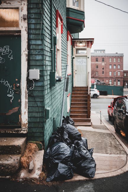 A small urban entrance area adjacent to a green wooden building is shown in the image, with a set of wooden stairs leading up to a white door with red trim. The building's exterior is clad in weathered green wooden siding with visible textures, and there are utility boxes and wiring attached to the wall. At the base of the steps, a pile of black rubbish bags is stacked on the concrete sidewalk, with some debris and a piece of brown burlap material nearby. In the background, a parking area is visible, with a few cars parked along the street and a larger brick building further away. The scene is set during daylight hours with overcast lighting, emphasizing the everyday context of private waste handling and alternative rubbish disposal arrangements, such as on-site collection by rubbish removal services like Rubbish Removal Knightsbridge, as opposed to council-provided waste collection.