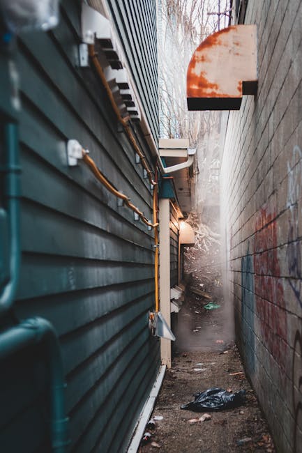 A narrow alleyway between two tall buildings with exterior walls, one clad in dark horizontal siding and the other in plain brickwork, both showing signs of weathering and graffiti. On the left side, a series of orange and black electrical cables and conduits are attached to the siding, running along the wall's surface. The ground is uneven and cluttered with scattered debris, including a crumpled plastic bag in the foreground. A faint warm light illuminates part of the alley from above, and in the background, there is a misty atmosphere with some bare tree branches visible against a partly cloudy sky. The scene suggests an area that might require private waste handling or rubbish removal, which Rubbish Removal Knightsbridge could address as part of an on-site clearance service or alternative waste collection method suited for tight urban spaces.