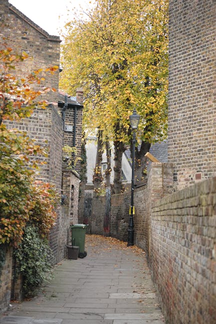 A small urban entrance area adjacent to a green wooden building is shown in the image, with a set of wooden stairs leading up to a white door with red trim. The building's exterior is clad in weathered green wooden siding with visible textures, and there are utility boxes and wiring attached to the wall. At the base of the steps, a pile of black rubbish bags is stacked on the concrete sidewalk, with some debris and a piece of brown burlap material nearby. In the background, a parking area is visible, with a few cars parked along the street and a larger brick building further away. The scene is set during daylight hours with overcast lighting, emphasizing the everyday context of private waste handling and alternative rubbish disposal arrangements, such as on-site collection by rubbish removal services like Rubbish Removal Knightsbridge, as opposed to council-provided waste collection.