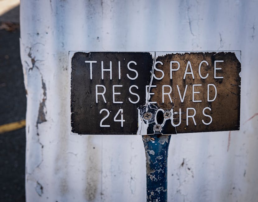 A weathered rectangular metal sign affixed to a pole, indicating a parking or reserved space. The sign has a black background with white, uppercase printed letters that read 'THIS SPACE RESERVED 24 HOURS.' The surface of the sign shows rust, peeling paint, and signs of aging, with parts of the material flaking away. The pole supporting the sign is painted blue, with visible chipped paint revealing patches of rust underneath. The background features a worn, white-painted wall or surface, with visible cracks and peeling paint, suggesting an outdoor or industrial environment. Natural daylight illuminates the scene, highlighting the textures of the rusted metal and weathered paint. This scene complements the context of alternative waste management or private property signage, as part of an environment where attention to property details and access rules is common, aligning with the themes of rubbish removal and site regulation.