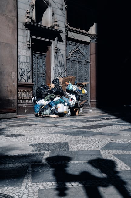 A large pile of mixed waste, including black plastic trash bags, cardboard boxes, plastic containers, and other discarded packaging, is stacked against the exterior wall of a historic building with ornate stone architecture and metal barred windows. The waste occupies a section of patterned pavement, with some shadows cast by nearby structures visible on the ground, indicating daylight conditions. The scene suggests an instance of unmanaged rubbish accumulation on a city street, which private waste removal services such as Rubbish Removal Knightsbridge may handle as part of alternative rubbish collection or on-site clearance. The building's detailed stonework and the graffiti marks on the lower walls provide context of an urban environment where rubbish may temporarily accumulate before removal. This image illustrates a typical scenario that requires professional waste management to maintain cleanliness in busy or historic city areas, emphasizing the importance of efficient rubbish disposal solutions beyond standard municipal collection services.