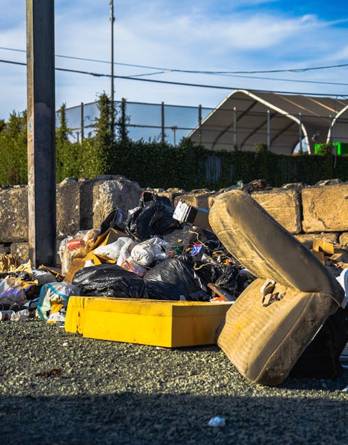 A pile of discarded rubbish situated outdoors on a gravel surface, consisting of black plastic trash bags, a yellow plastic container, a worn beige vehicle bumper, and various loose debris. The waste appears to include packaging materials and miscellaneous refuse. Behind the pile, there is a low stone wall made of rectangular blocks, with some greenery and small trees visible beyond it. To the left, a metal utility pole with power lines extends upward, while in the background, a partially visible structure with a curved roof can be seen under a bright blue sky with some light clouds. The scene suggests an area where waste has been temporarily dumped, potentially related to private waste clearance services such as those provided by Rubbish Removal Knightsbridge, reflecting an informal or non-licensed collection site.