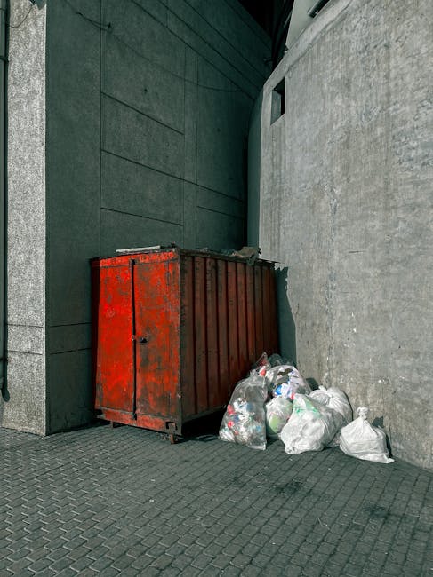 A red metal skip with a weathered, slightly rusted surface is positioned against the corner of a building's exterior wall, which is composed of dark green panels and light grey concrete. The skip is partially obstructed by several clear and opaque plastic bags filled with mixed rubbish, including crumpled paper, plastic bottles, and miscellaneous waste materials, situated directly on the paved ground made of dark grey interlocking bricks. The scene is set outdoors, in an urban environment, with the skip placed in a narrow alleyway or side passage beside the building. The lighting suggests natural daylight, casting soft shadows and highlighting the textures of the materials and surfaces. This setting reflects typical private waste disposal arrangements, where independent rubbish collection can be managed through external skips, distinct from municipal collection services, aligning with waste management practices like those offered by Rubbish Removal Knightsbridge.