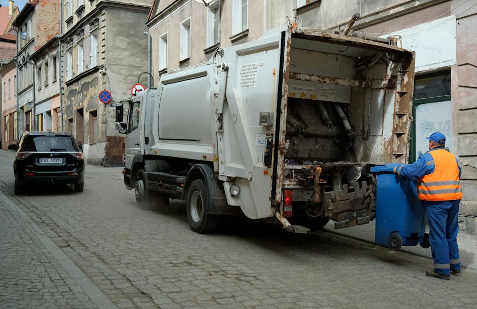 A worker wearing a blue uniform and a high-visibility orange vest is emptying a small blue trash bin into the rear compartment of a white waste collection truck parked on a narrow cobblestone street in an urban area. The truck's rear door is open, revealing a rusty and worn interior designed for compacting household or commercial rubbish. The street is lined with old-style multi-story buildings with stone and brick facades, some showing signs of weathering and peeling paint. A dark-colored car is parked further down the street beside the curb, and street signs are visible on the building wall, indicating parking restrictions. Natural daylight illuminates the scene, highlighting the textures of the cobblestones, the metallic surfaces of the waste truck, and the fabric of the worker's uniform. The environment suggests an area where private waste handling and independent collection methods are employed, in line with local rubbish removal services, such as those provided by Rubbish Removal Knightsbridge.