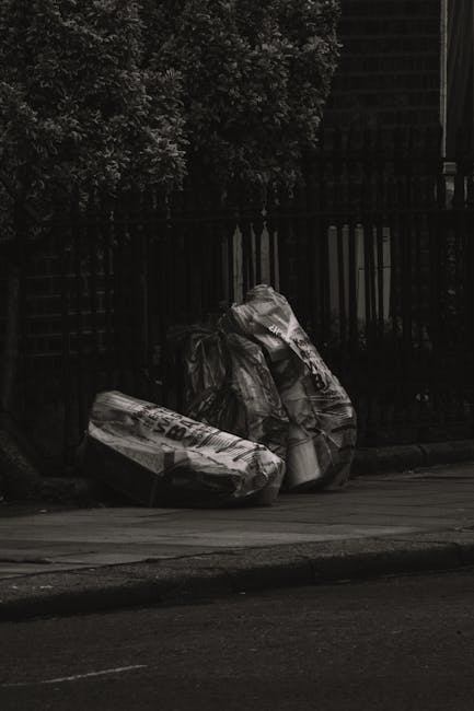 In the foreground, there are several large plastic bags filled with waste, positioned on the side of a paved street or sidewalk. The bags appear to be crumpled and loosely tied, with some partially spilling contents that suggest mixed rubbish material. Behind the bags, there is a black metal fence with vertical bars, separating the waste from a background area that includes dense foliage from trees or bushes, which adds a natural element to the scene. The environment is outdoors with minimal lighting, giving the image a somewhat shaded or subdued appearance, indicating it might have been taken during overcast conditions or in low light. The surface of the pavement shows signs of wear and is clean from other debris. The presence of the waste bags on the street edge implies an intermediate stage of rubbish collection, typical of private waste disposal or independent clearance services, as managed by organisations such as Rubbish Removal Knightsbridge. Overall, the scene reflects a typical urban setting where waste is temporarily stored outside prior to collection or removal, with a focus on the visual clarity of the bags and surrounding environment, suitable for content related to rubbish removal services and alternative waste handling options.