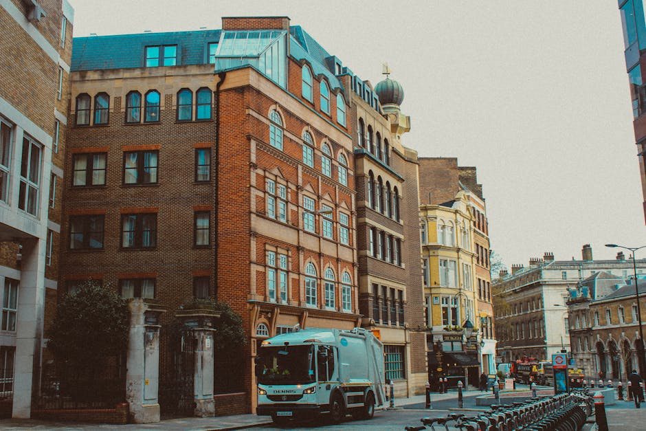 A city street scene featuring a multi-story brick building with ornate window architraves and a rounded turret topped with a domed roof, situated among other commercial and residential structures. In the foreground, a light blue waste collection vehicle with a compact, modern design is parked on the pavement, positioned slightly to the right. The vehicle's surface is smooth, with visible windows and a compact cargo area. To the left of the vehicle, there is a stone entrance gate with two pillars and foliage nearby. The street is lined with metal bike racks, some of which are empty, and bollards separating the pedestrian walkway from the roadway. The environment appears to be an urban area with a mix of historical and contemporary architecture, under overcast lighting that creates a neutral, professional atmosphere typical of services like rubbish removal, supporting private waste management efforts near Knightsbridge.