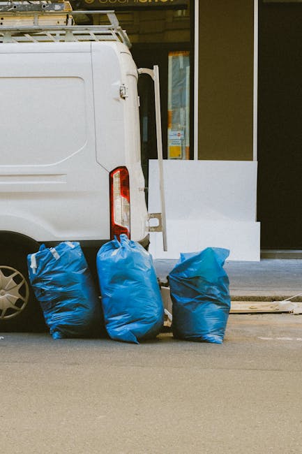 Close-up view of the rear side of a white commercial van parked on a city street, with three blue rubbish bags leaning against the lower part of the vehicle near the left rear wheel. The bags appear to be filled with waste, with crinkled plastic surfaces and sealed tops. The background features a portion of a building with a glass door and window, displaying some signage and lighting inside, along with a white step or barrier setting off the entrance area. The scene suggests an on-site waste collection or private rubbish removal service, typical of what Rubbish Removal Knightsbridge may provide for local clients seeking alternative disposal options outside traditional council collections. The asphalt pavement has a neutral grey tone, and the lighting indicates daytime with natural ambient light, contributing to a clear and unobstructed view of the rubbish bags and the vehicle, all within an urban environment dedicated to waste management similar to services outlined on the page about weekend rubbish pickup in Knightsbridge.