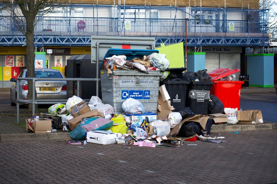 A close-up view of a large pile of mixed waste and rubbish situated on a gravel surface outdoors. The collection includes black plastic trash bags, a torn cardboard box, an old, worn car tire leaning against the rubbish, and assorted discarded materials. Behind the waste are rough, beige-colored stone blocks arranged as a barrier or retaining wall, with some greenery and bushes visible beyond. In the background, there is a metal fence, utility poles with wires, and a partially visible large structure with a curved roof, possibly a sports or storage facility, under a partly cloudy sky illuminated by natural daylight. The scene reflects a typical area for private waste disposal, possibly managed by independent rubbish removal services, such as Rubbish Removal Knightsbridge, highlighting the importance of professional rubbish collection and on-site clearance for environmentally responsible waste management.